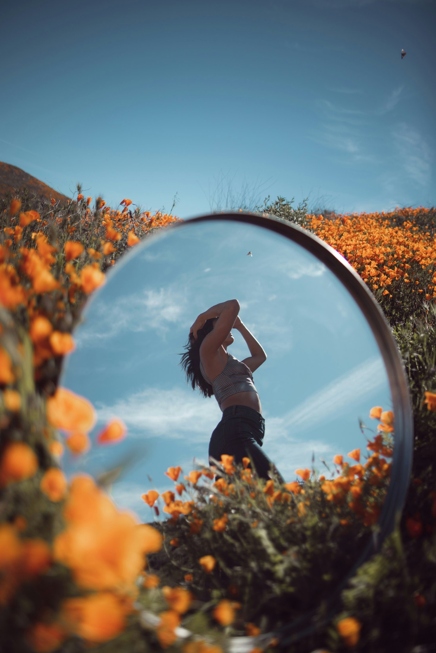 Un espejo entre flores naranjas refleja a una mujer bajo el cielo