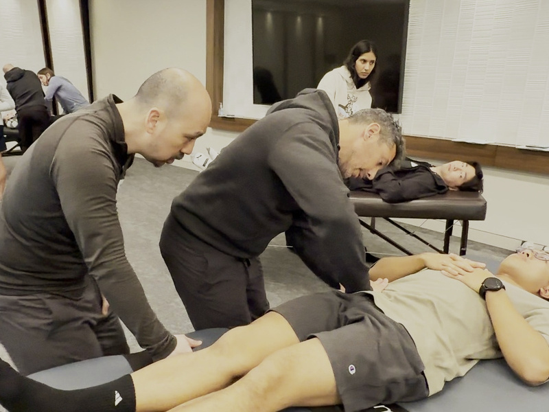 Two instructors working through a leg technique on a student at the table