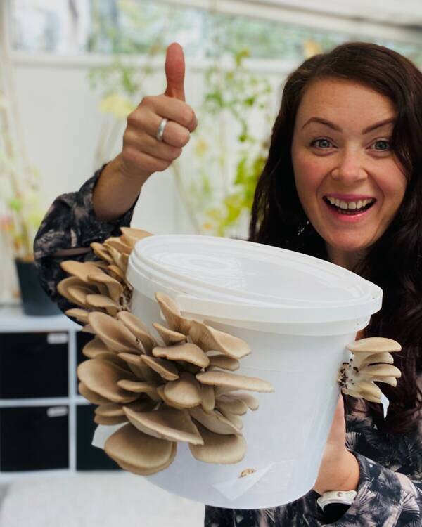 A woman grinning and giving a thumbs-up, holding a bucket sprouting tan oyster mushrooms