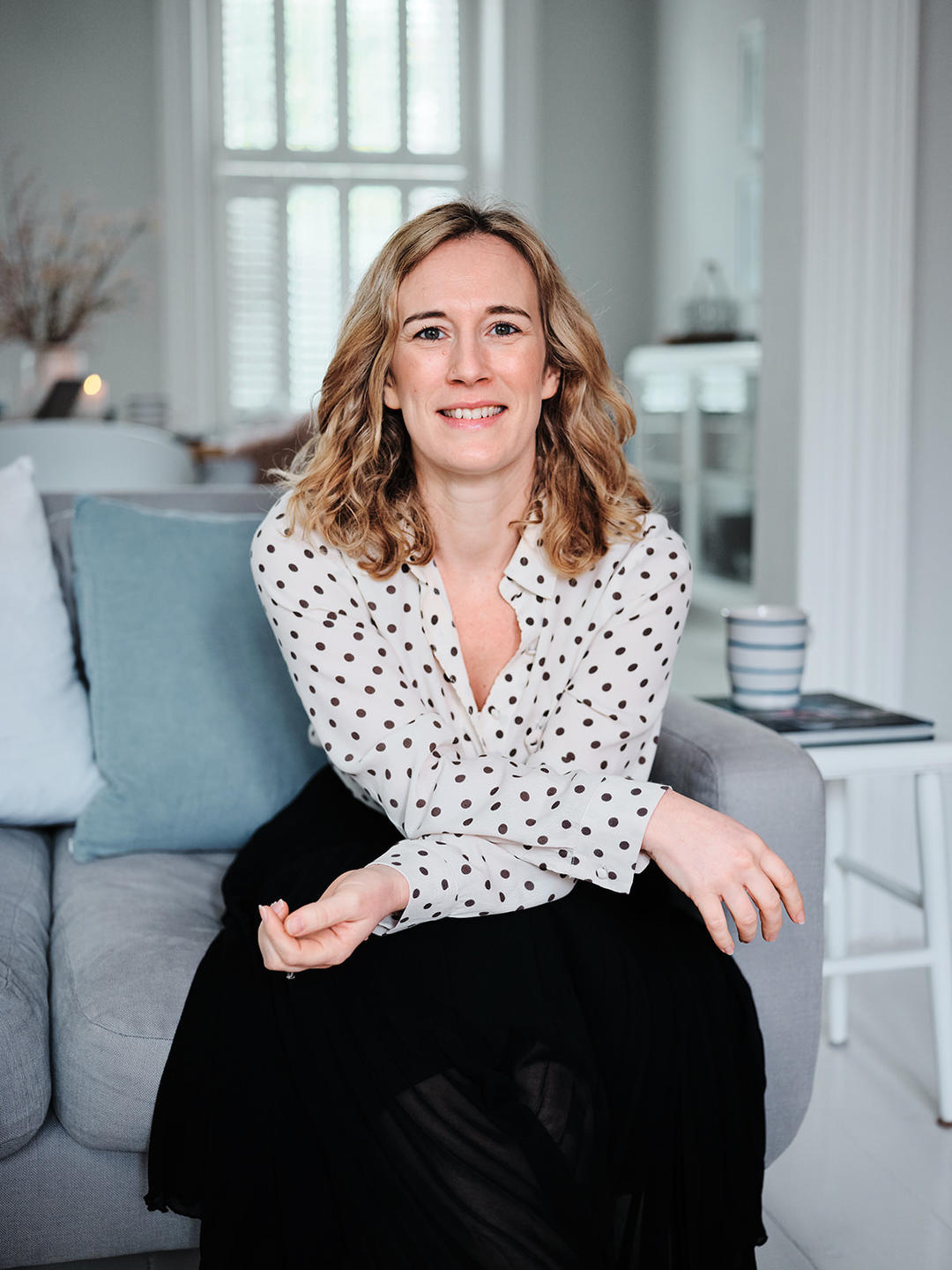 Picture of Zoe Hawkins in a spotted blouse and black skirt, sat in a chair, smiling, with her arms crossed leaning forwards