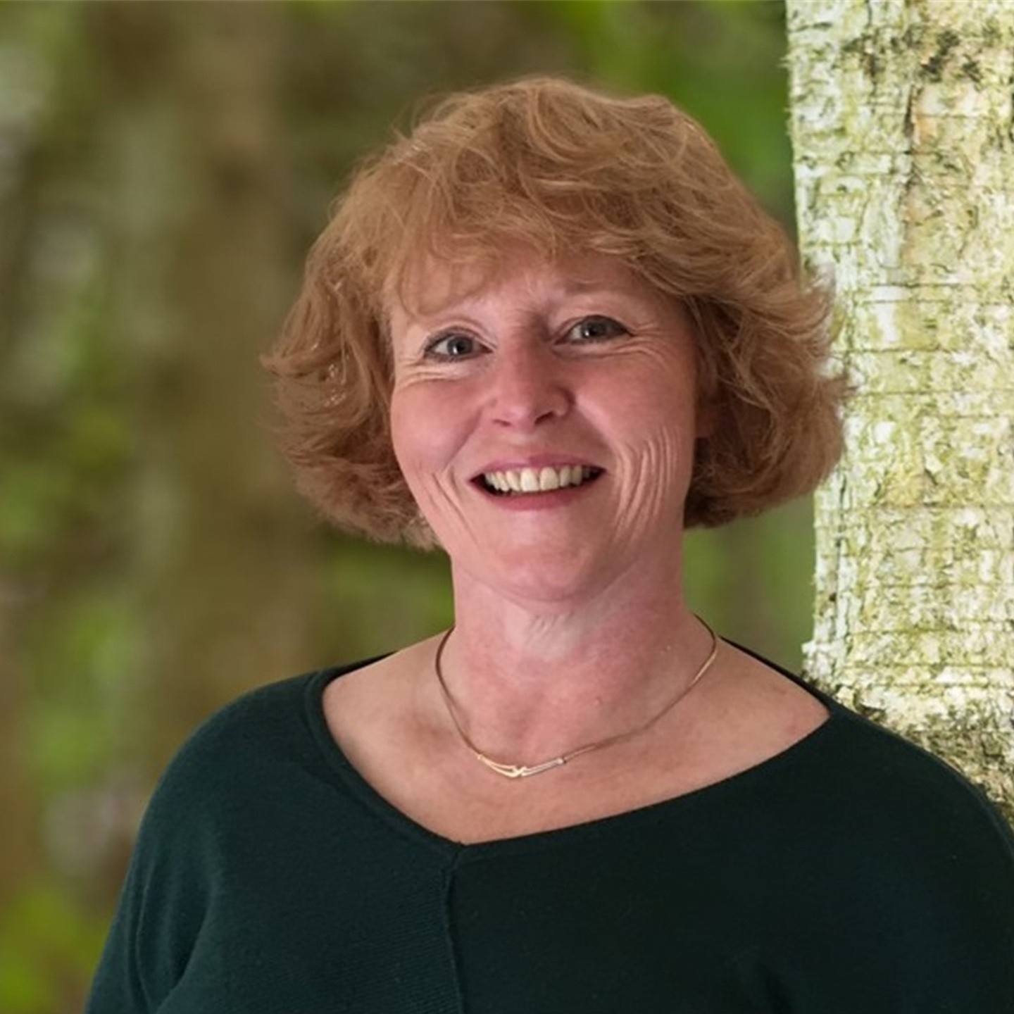 Photo of Helen Harding, a 50 year old smiling woman with mid lenght red hair wearing a green jumper standing in front of a tree