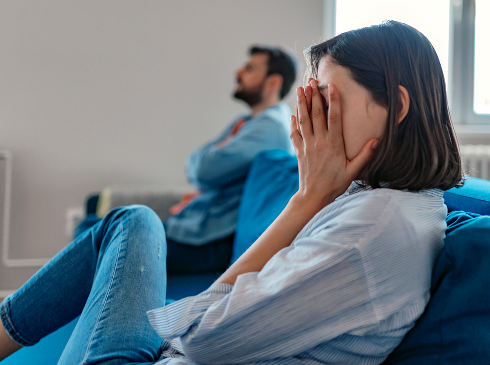 Woman sitting on opposite end of sofa from partner with her face in her hands